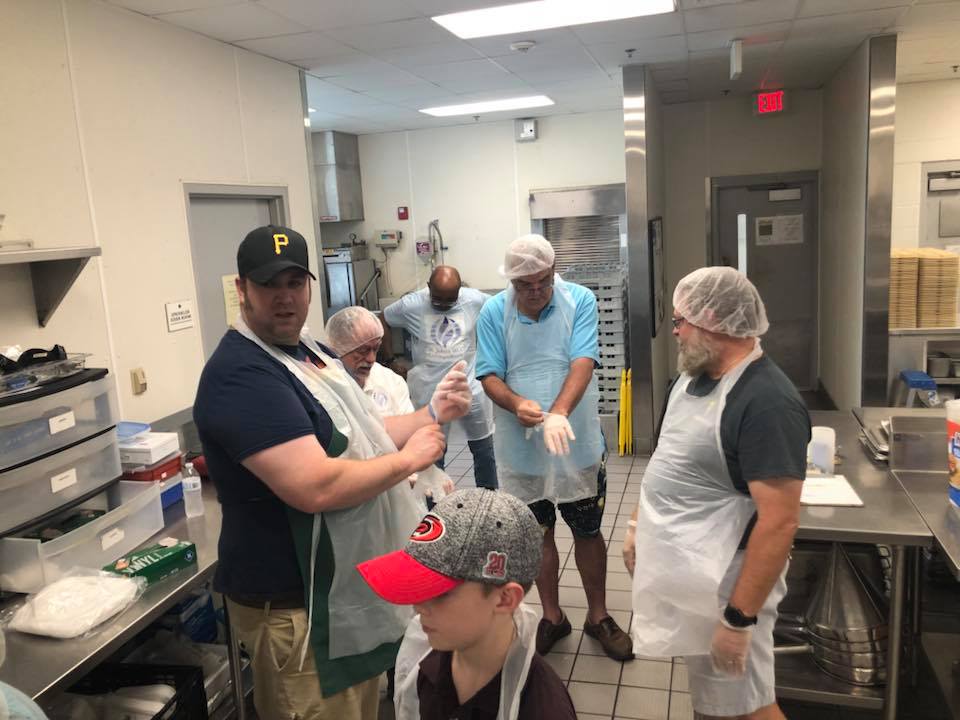 A group of 5 volunteers from St. John's MCC standing in a kitchen putting on work gloves and wearing aprons preparing to serve lunch boxes of love in Raleigh North Carolina